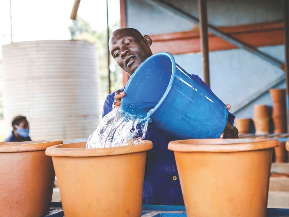 A person pours water from a blue bucket into large clay pots. They appear focused, with a workshop and stacked pots in the background, suggesting pottery work.