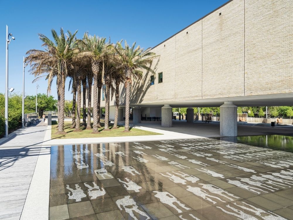 Modern building with a white brick facade and palm trees reflected in a shallow pool with human-shaped outlines.