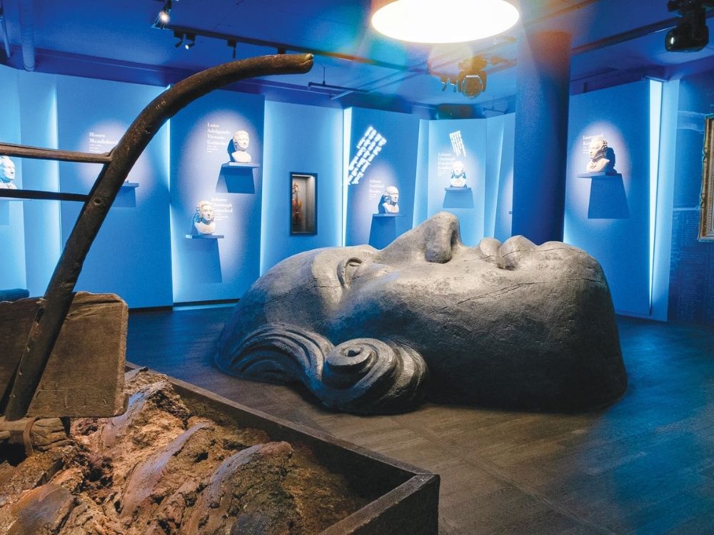 A dimly lit museum room features a large, reclining stone face sculpture and busts on blue-lit walls.