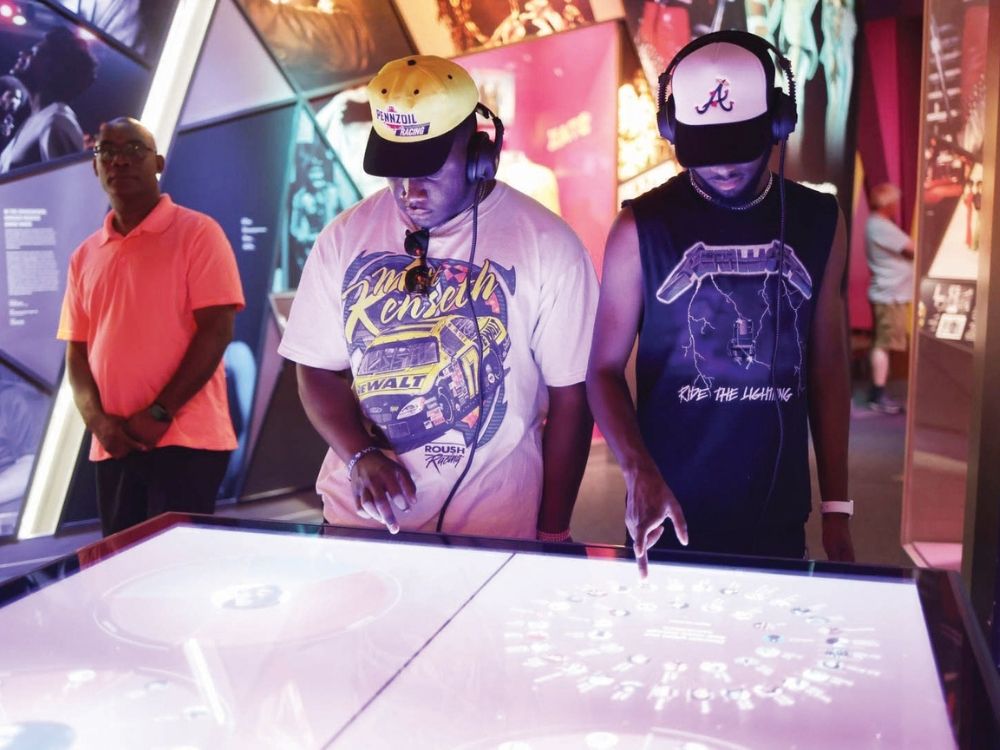 Two men wearing caps and headphones interact with a digital touchscreen display at a museum exhibit. Another man observes in the background.