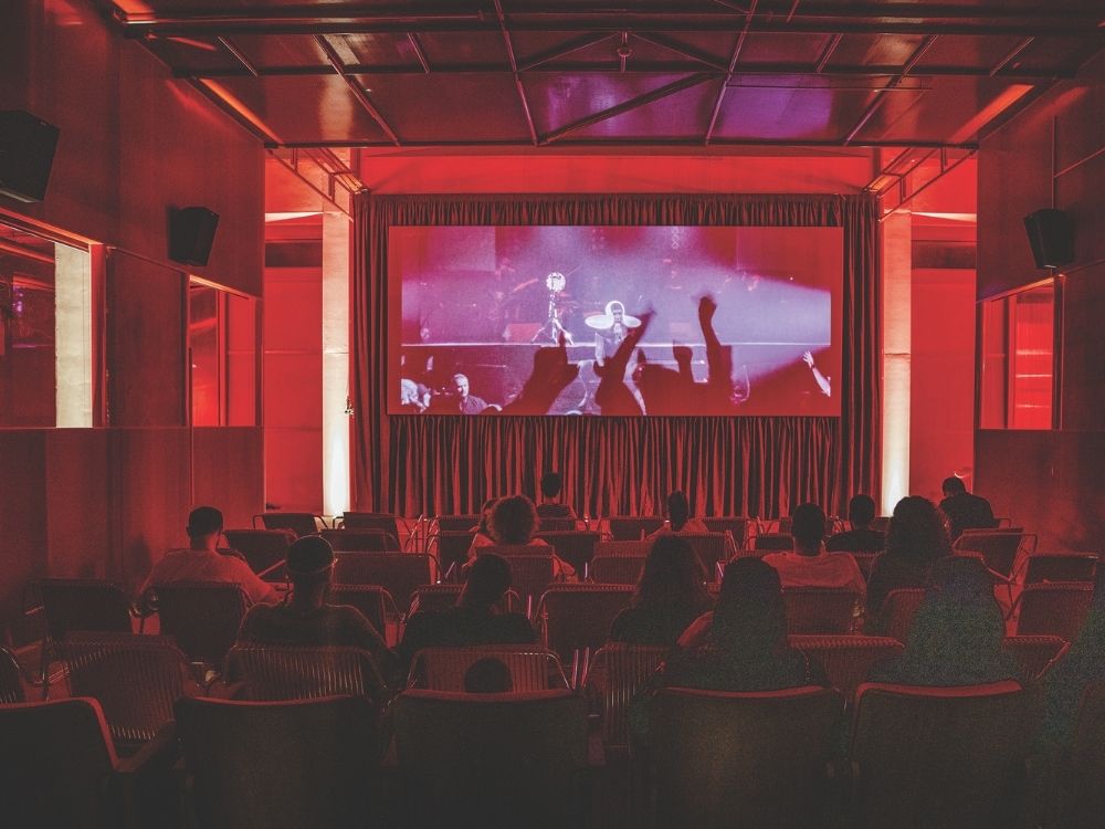 A dimly lit theater with people seated, watching a large screen at the front. The room is bathed in red light, creating an immersive atmosphere. Audience shadows are visible on the screen.