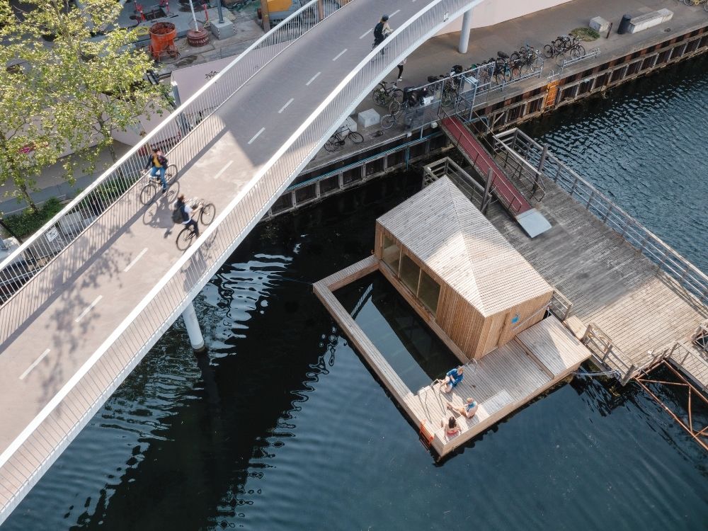 Aerial view of a modern bridge with cyclists above a calm canal. Below, a wooden floating structure with people relaxing on the deck.