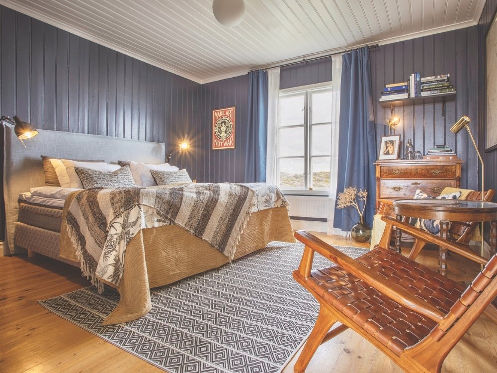 Cozy bedroom with dark blue paneled walls, a large bed with patterned throw, wooden floor, vintage furniture, and soft lighting from wall lamps.