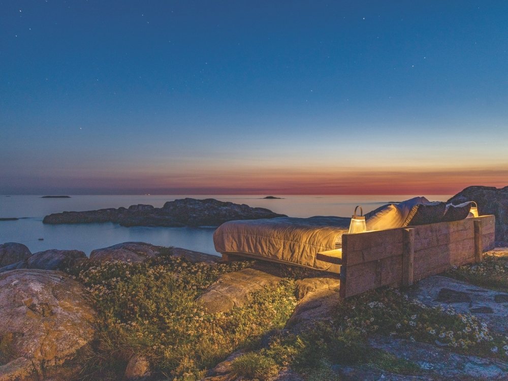 A cozy wooden bed with soft bedding is placed on rocky terrain, overlooking a tranquil ocean at sunset. The sky transitions from warm orange to deep blue.