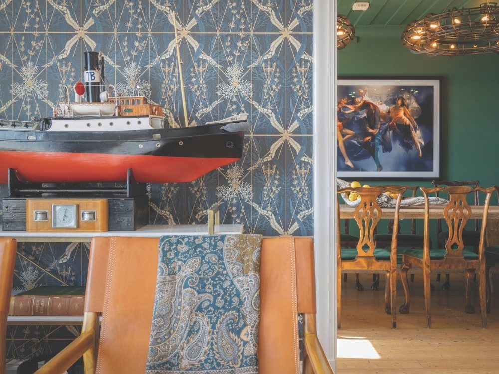 Vintage nautical-themed room with a model ship on a shelf, leather chairs, ornate wallpaper, and a vibrant underwater art piece in the background. Cozy and eclectic.