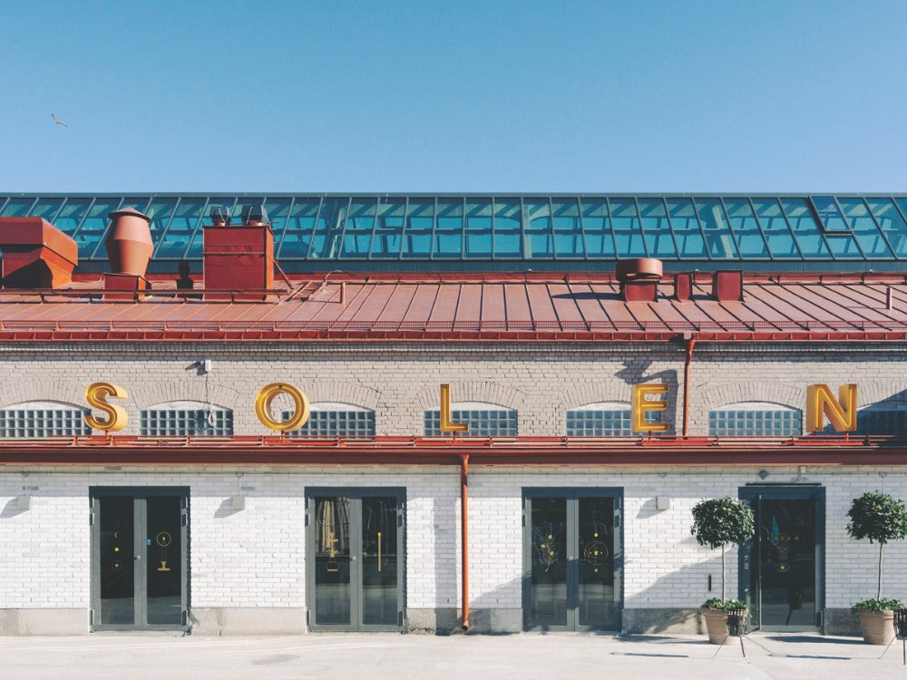 Historic brick building with a red roof and large yellow letters spelling "SOLEN." Blue sky above, green plants at the entrance. Calm, sunny ambiance.