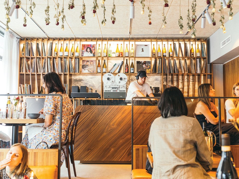 Modern cafe scene with dried flowers hanging from the ceiling, a wooden counter with vinyl records and bottles, a DJ playing music, and relaxed patrons.