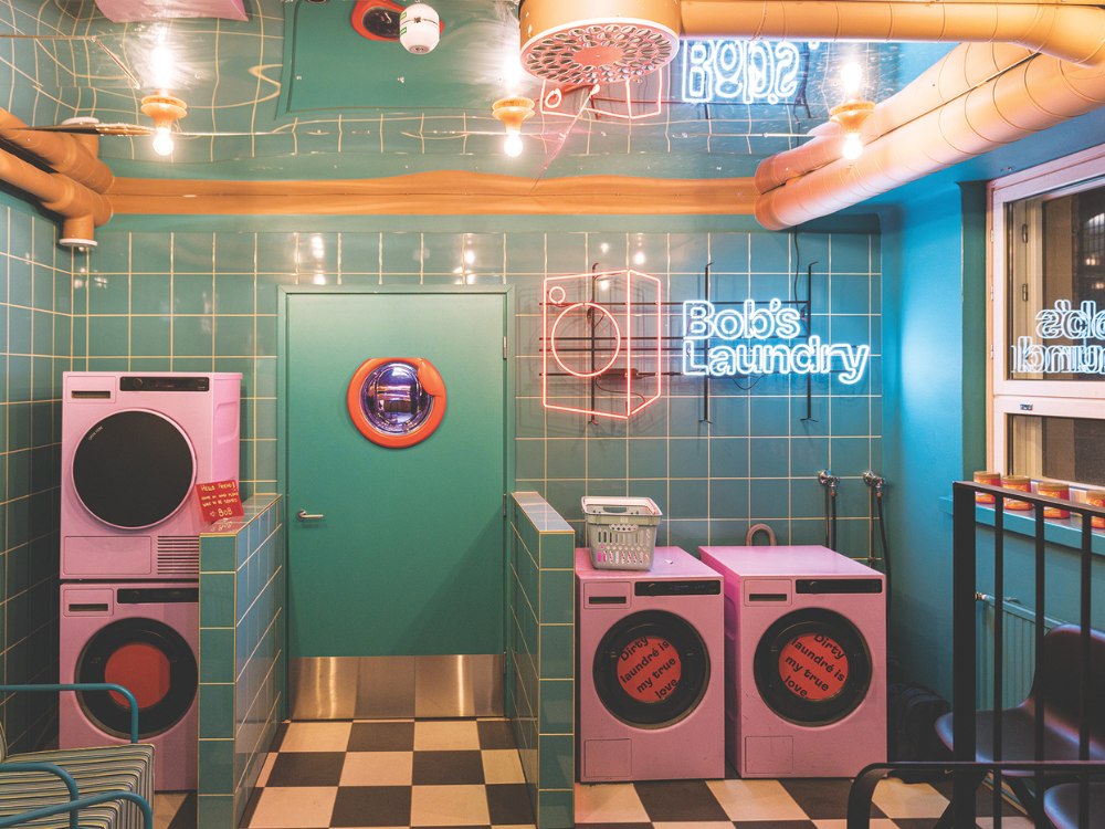 Laundry room with retro style, featuring pink washing machines and turquoise tiles. Neon signs read "Bob’s Laundry." Bright and playful ambiance.
