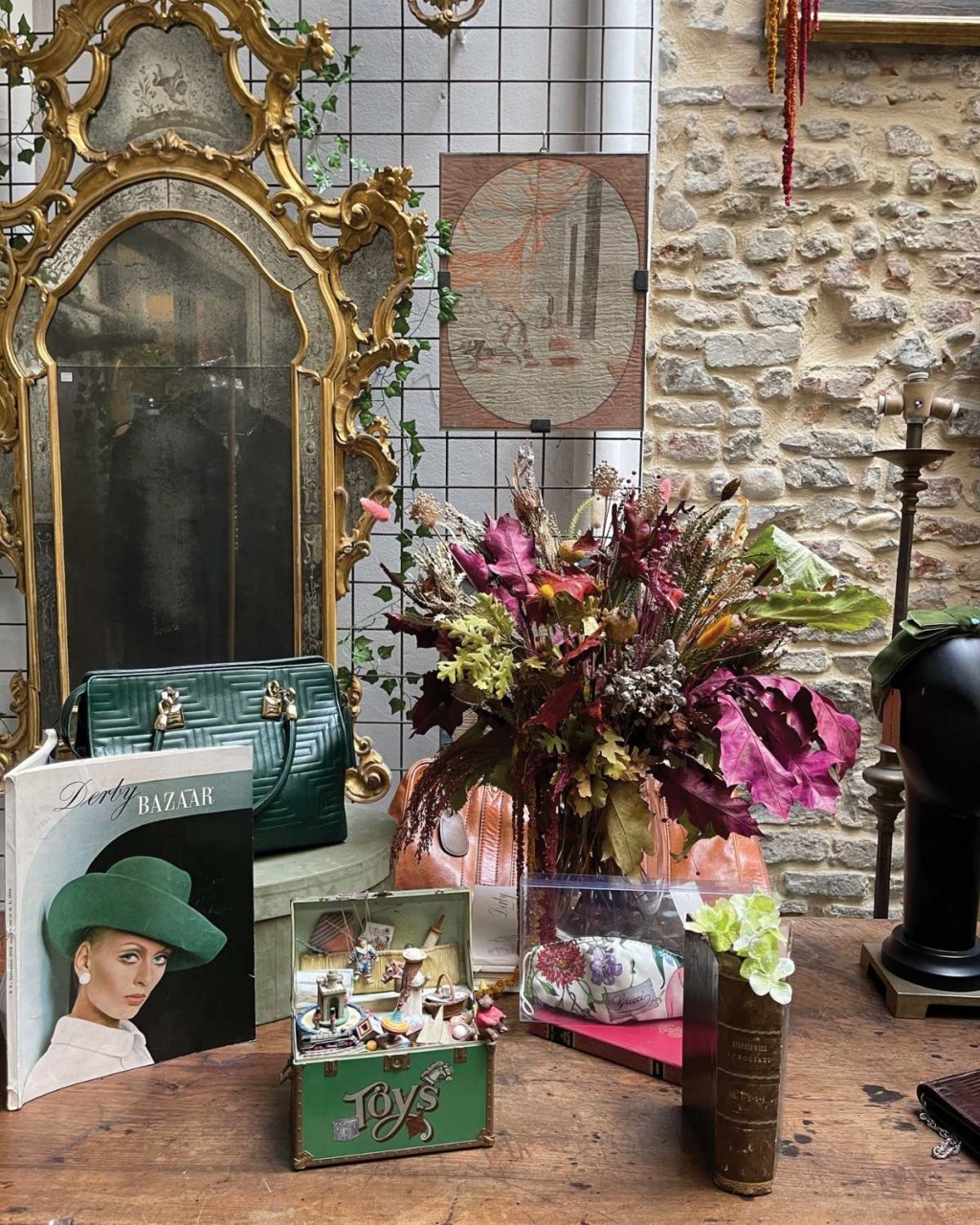 A vintage-style display featuring a large ornate mirror, a green handbag, a bouquet of dried flowers, and a collection of decorative items.