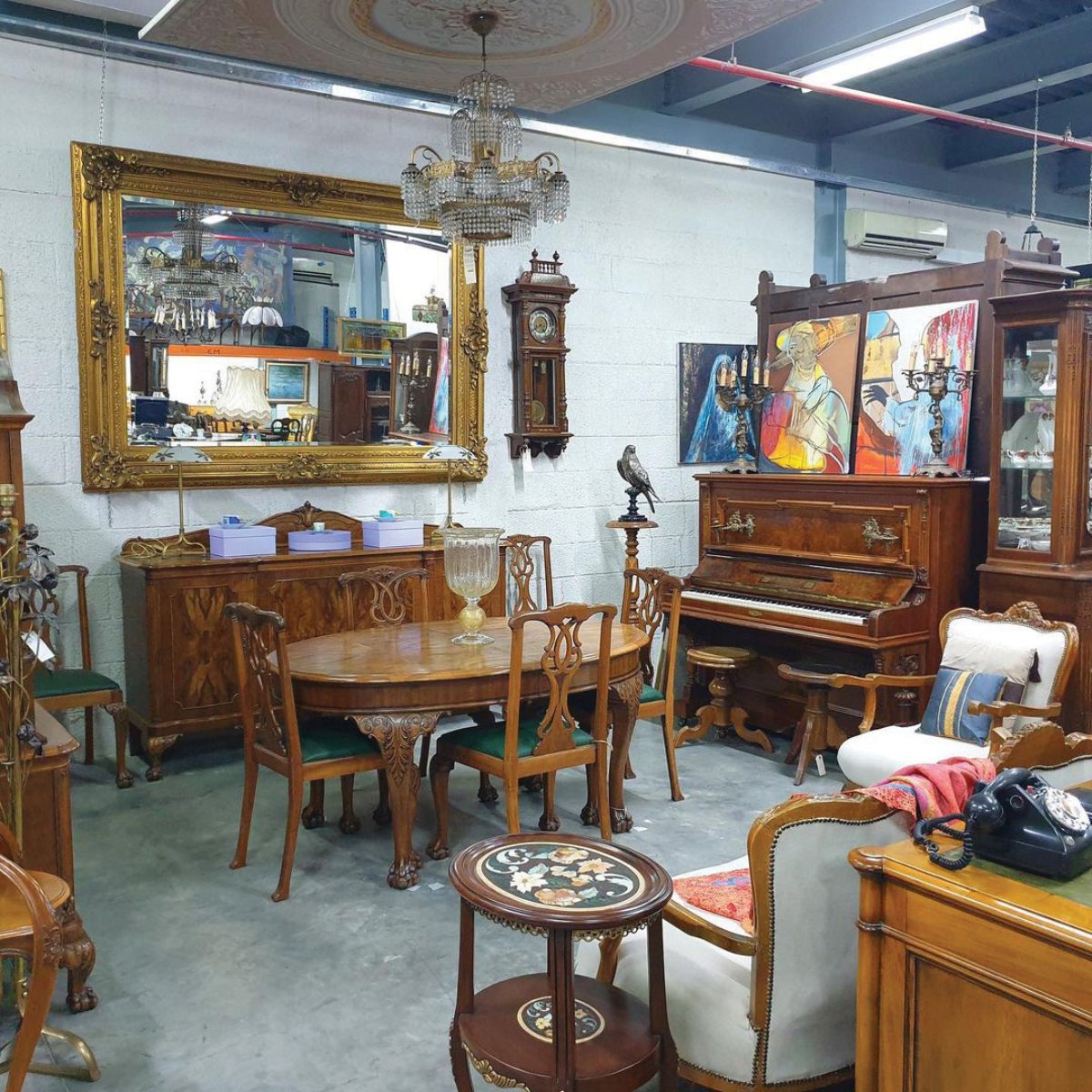A vintage furniture showroom featuring ornate wooden pieces, a grand mirror, a chandelier, and eclectic artwork on the walls.
