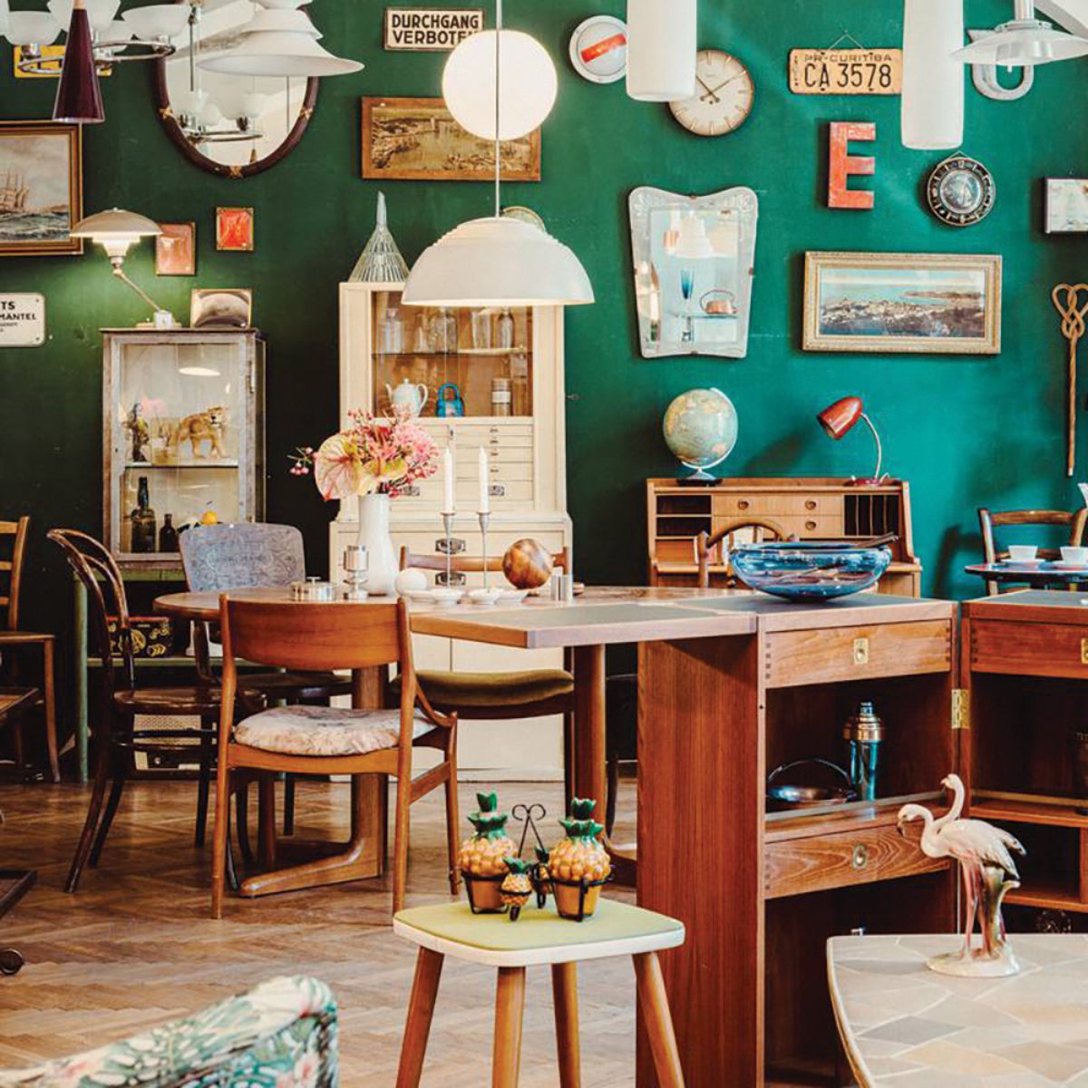 Vintage-themed room with a green wall adorned with eclectic decor, including framed art, clocks, and signs. Wooden furniture and retro items create a cozy atmosphere.