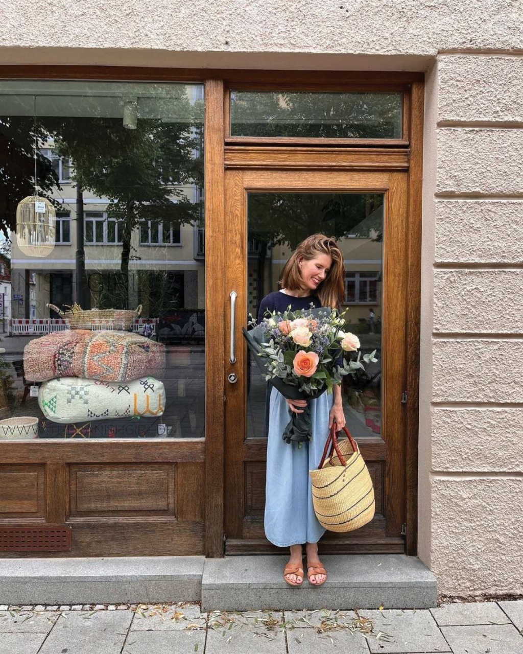 A person stands in front of a shop door, holding a large bouquet of flowers and a straw bag, with decorative blankets visible inside.
