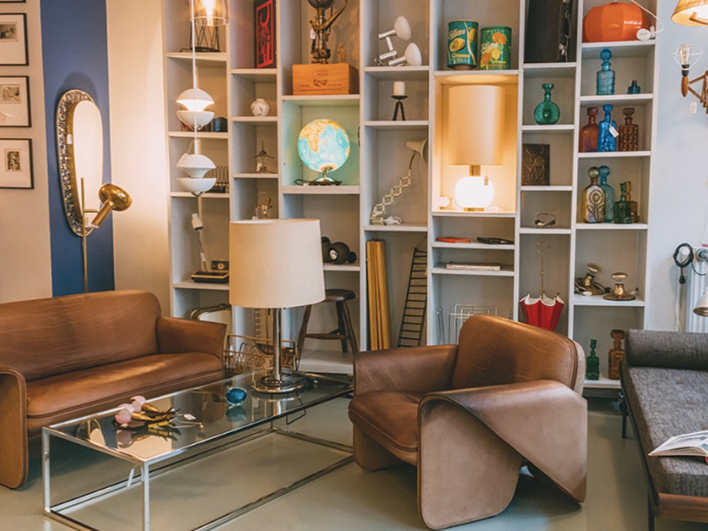 Cozy living room with vintage decor. Brown leather sofas, glass coffee table, and a tall lamp. Shelves display a globe, books, and colorful vases. Warm ambiance.