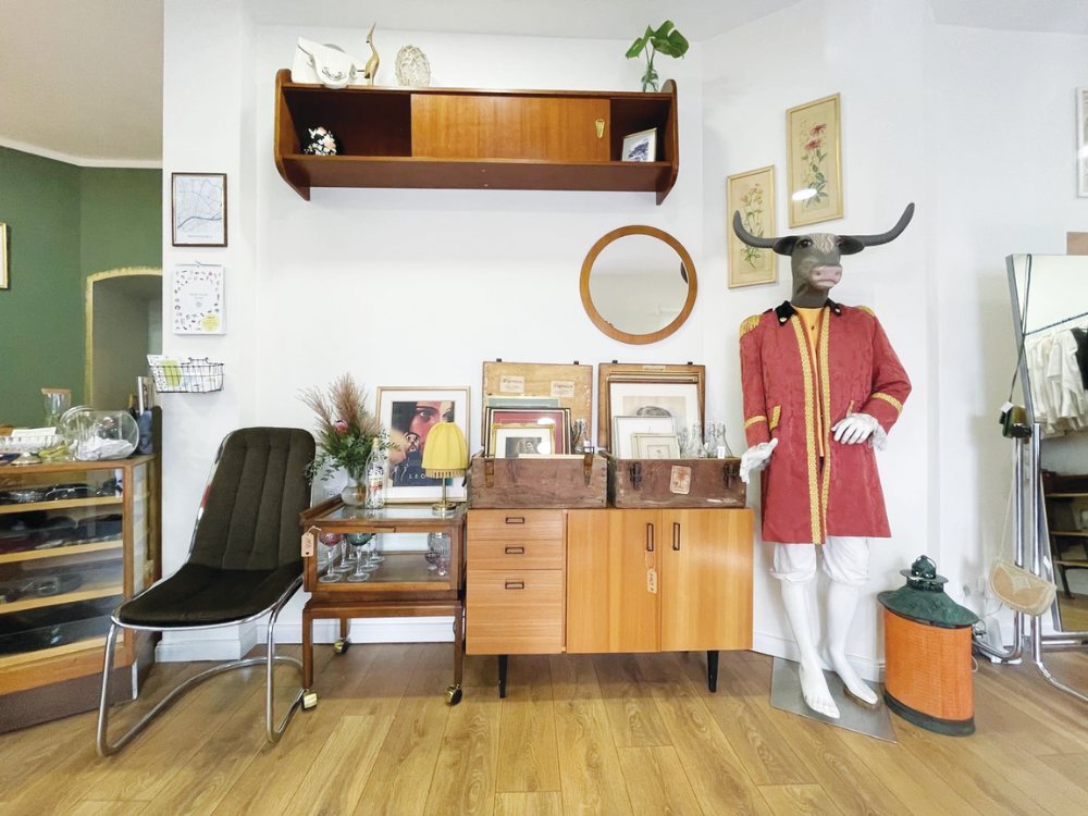 Vintage-style room with wooden furniture, a mannequin in a red coat with a bull head, framed art, and a mirror. The atmosphere is eclectic and retro.