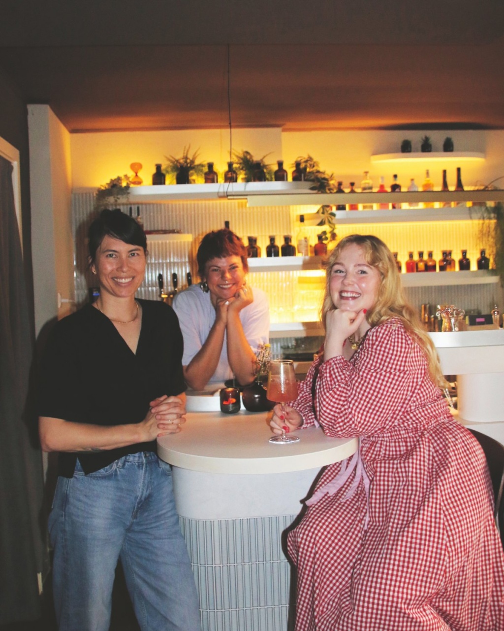 Three people smiling at a cozy bar with warm lighting. Shelves with bottles and plants decorate the background, creating a relaxed, friendly atmosphere.
