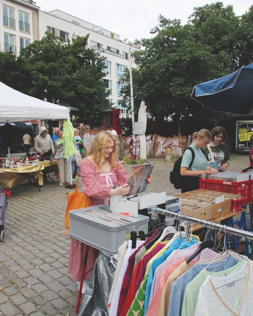 A woman in a red checkered dress browses records at an outdoor market. Nearby, racks display colorful clothes. The atmosphere is lively and casual.