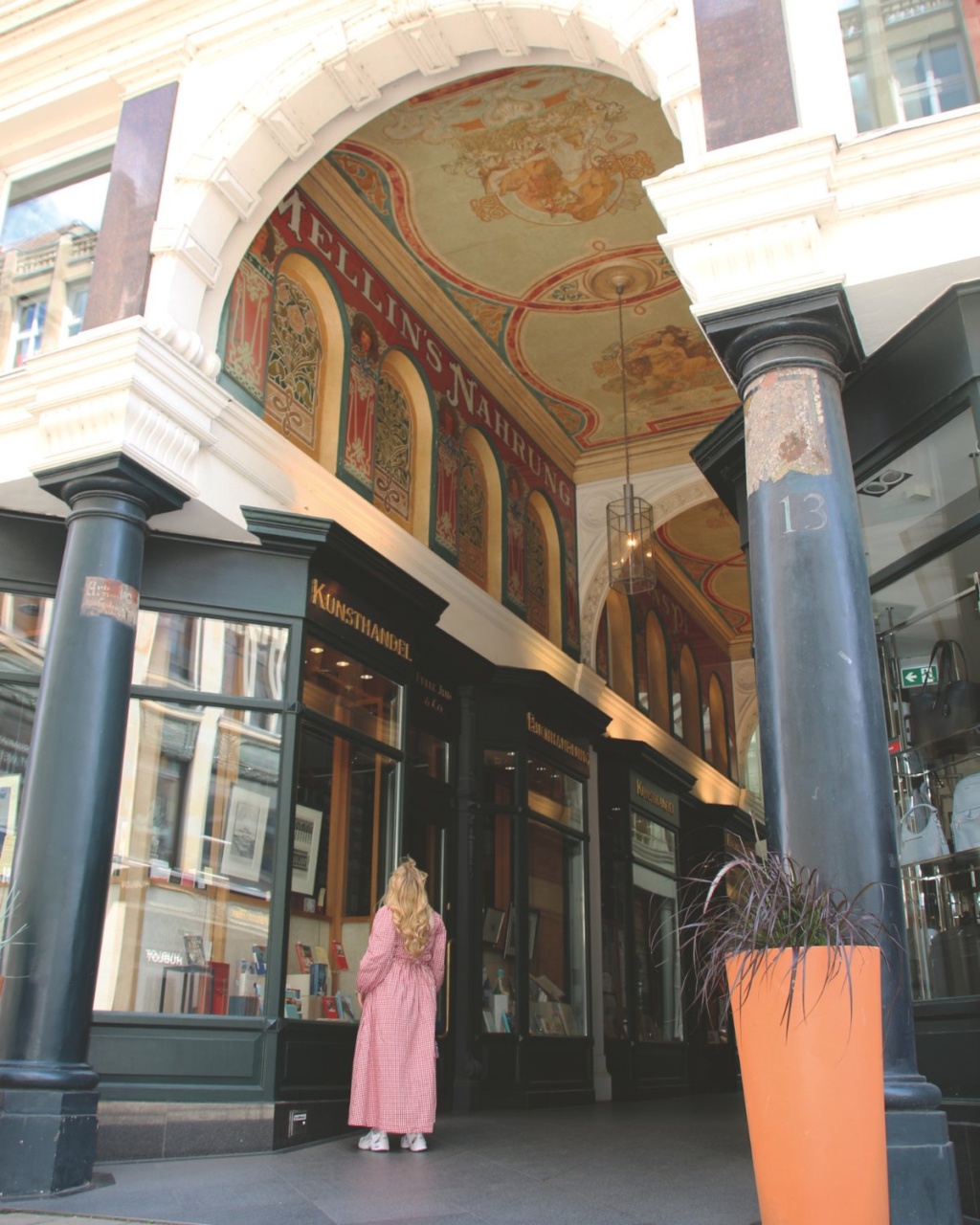 A person in a dress stands in an ornate arcade, gazing at shop displays. The ceiling features elaborate frescoes with warm tones, conveying a historic elegance.