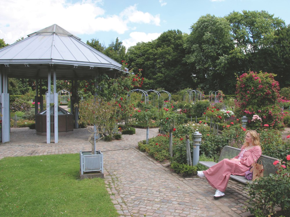 A woman in a red gingham dress sits on a bench in a lush rose garden, near a large gazebo. The scene is serene, with blooming flowers and green trees under a partly cloudy sky.
