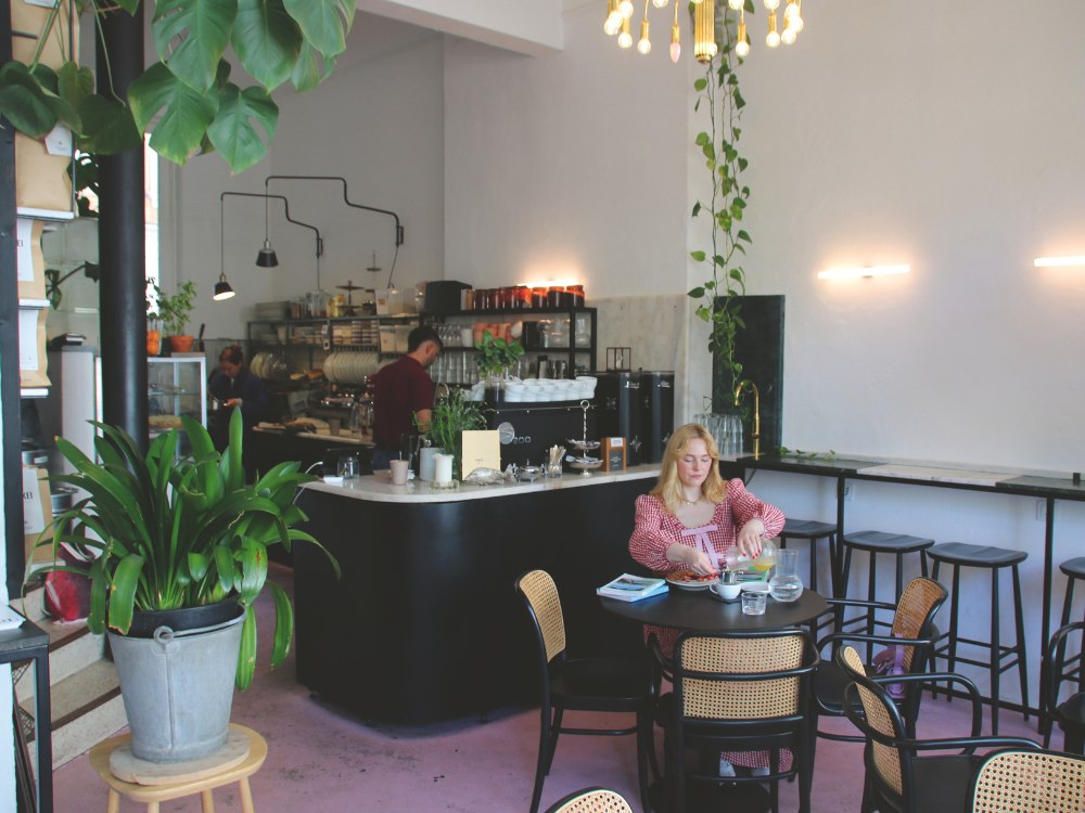 Cozy cafe with pink floors and plants, featuring a woman in a pink dress dining alone at a round table. In the background, a barista works behind a counter.