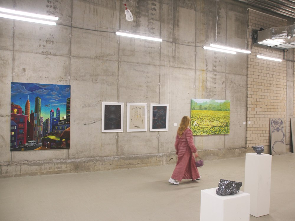 A woman in a red dress walks in an art gallery with concrete walls. Colorful cityscape and landscape paintings hang on the walls, with sculptures on white pedestals.