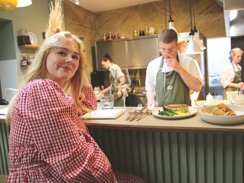 A woman in a red checkered dress sits at a restaurant counter, smiling. Chefs in green aprons prepare food, creating a cozy and inviting atmosphere.