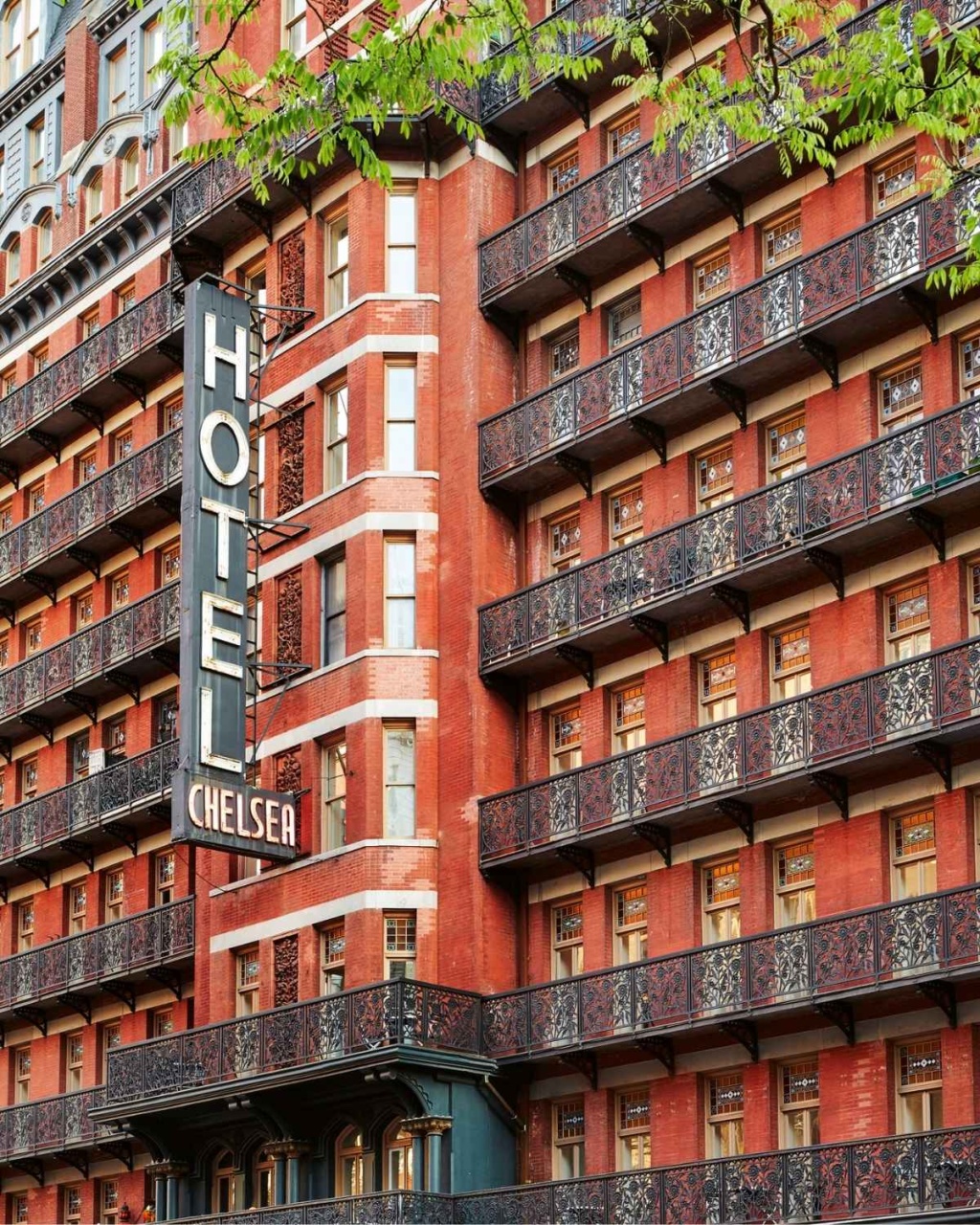 Red brick façade of a historic hotel with ornate black balconies and a prominent vintage sign reading "Hotel Chelsea".