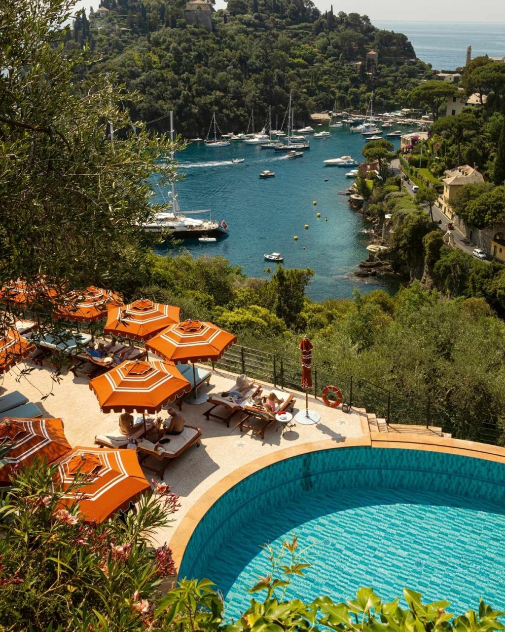 View of a tranquil bay with sailboats, surrounded by lush greenery. Foreground shows a curved pool, orange umbrellas, and sun loungers.