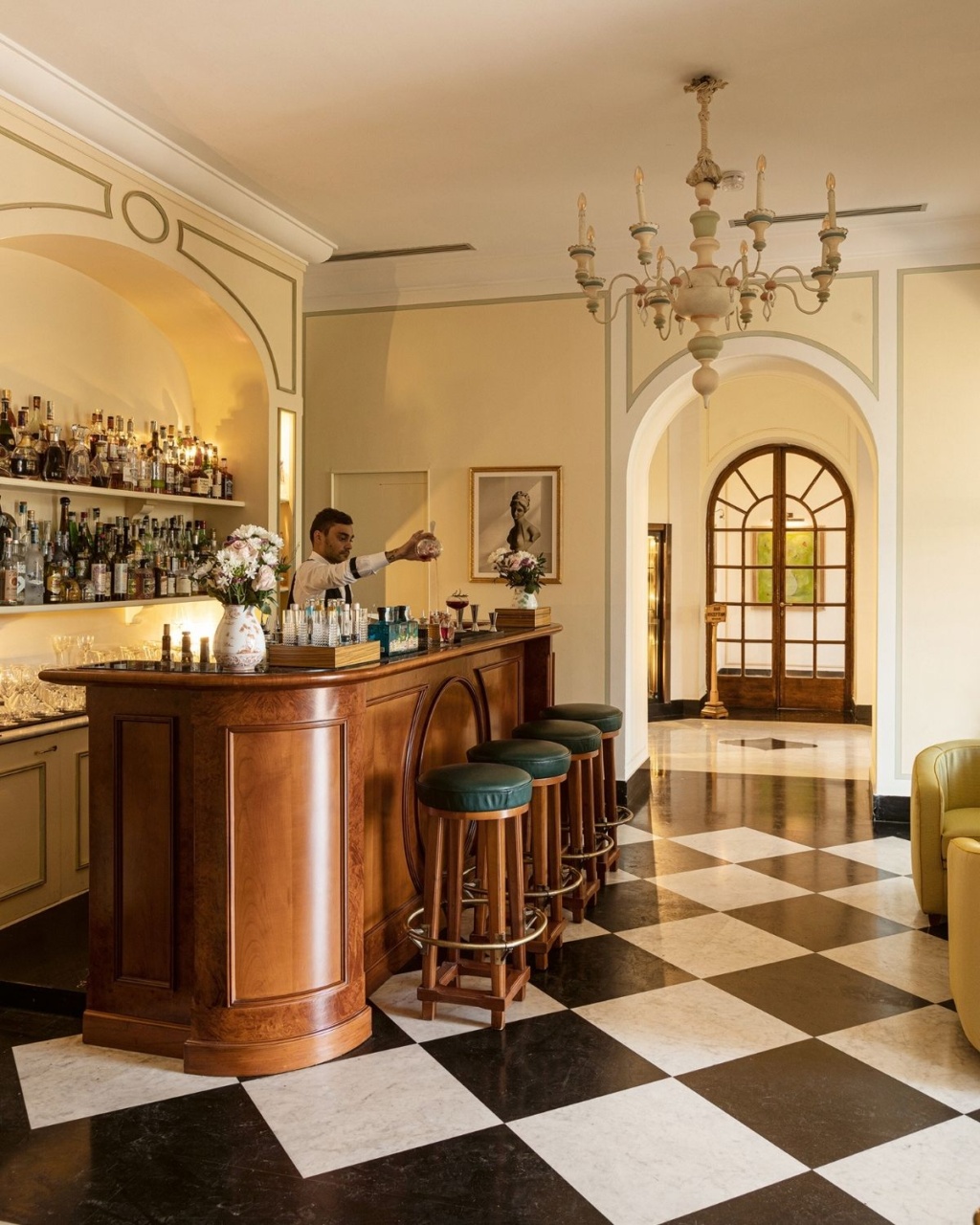 Elegant bar interior with a wooden counter, green stools, and checkered floor tiles. A bartender prepares drinks.