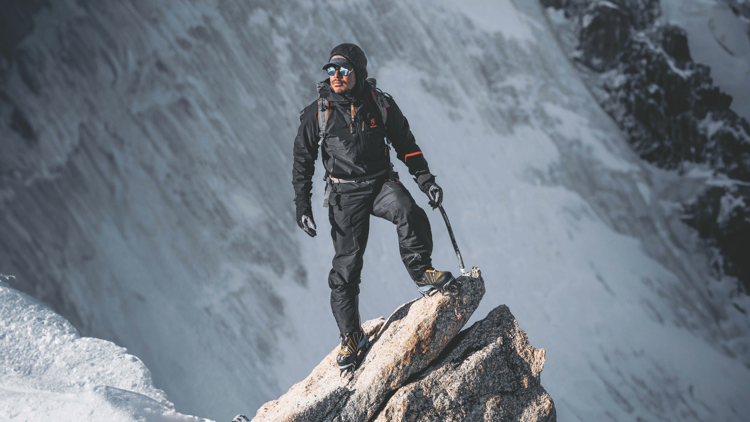 A climber in black gear stands confidently on a snowy mountain peak, holding an ice axe. The background features vast snowy slopes and rugged terrain.