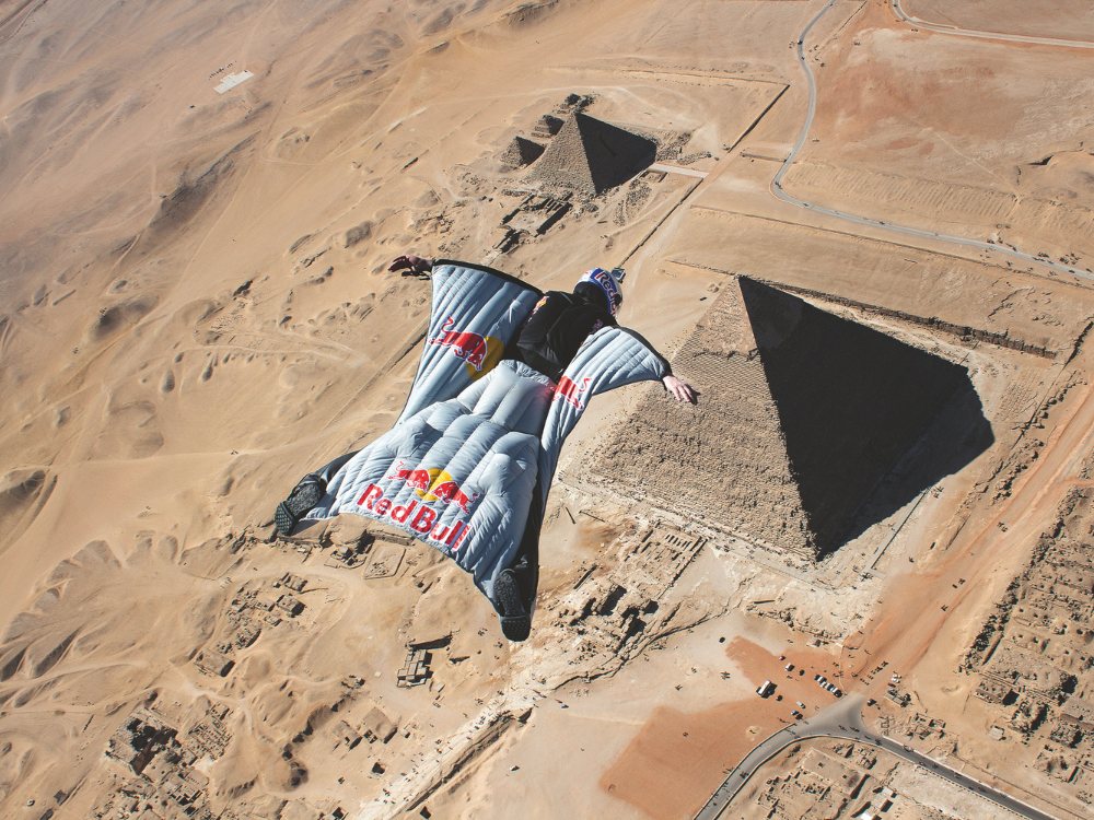 A person in a Red Bull wingsuit glides over the Pyramids of Giza in a desert landscape, conveying excitement and adventure from a bird's-eye view.