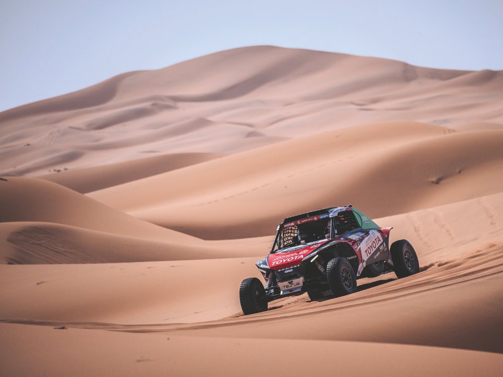 A rally car races across vast desert sand dunes, kicking up dust. The scene is dynamic and adventurous, with clear blue skies overhead.