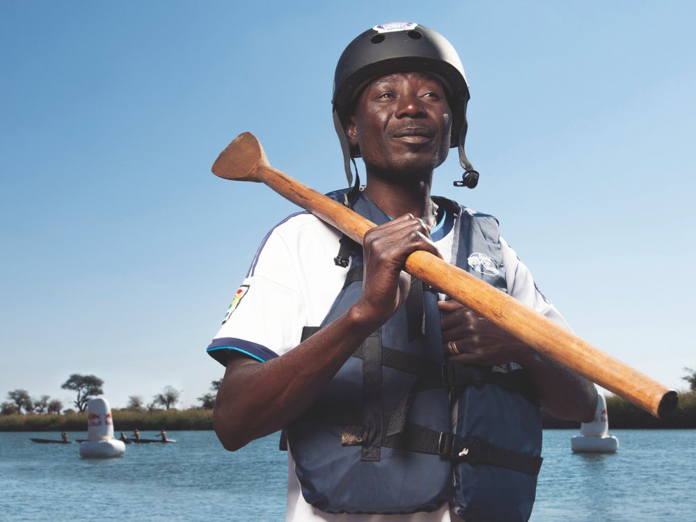 A man stands by a calm river, wearing a helmet and life vest, holding a wooden paddle confidently. The sky is clear, with trees in the background.