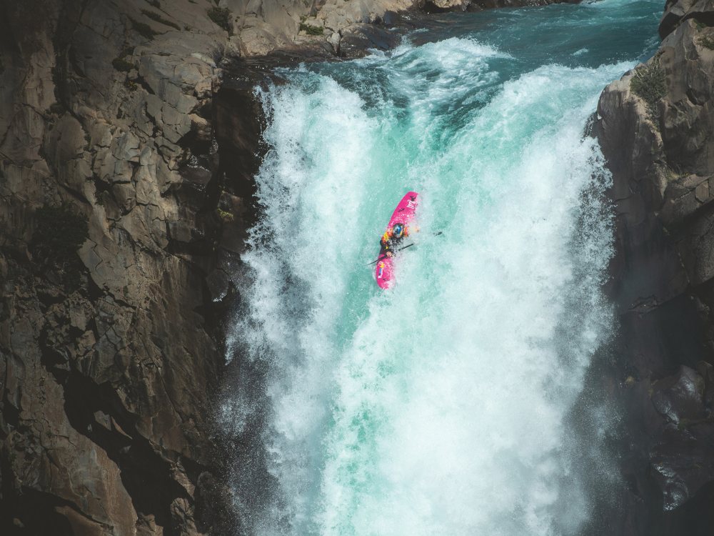 A kayaker in a bright pink kayak navigates down a powerful, turquoise waterfall flanked by rocky cliffs, conveying a sense of adventure and thrill.