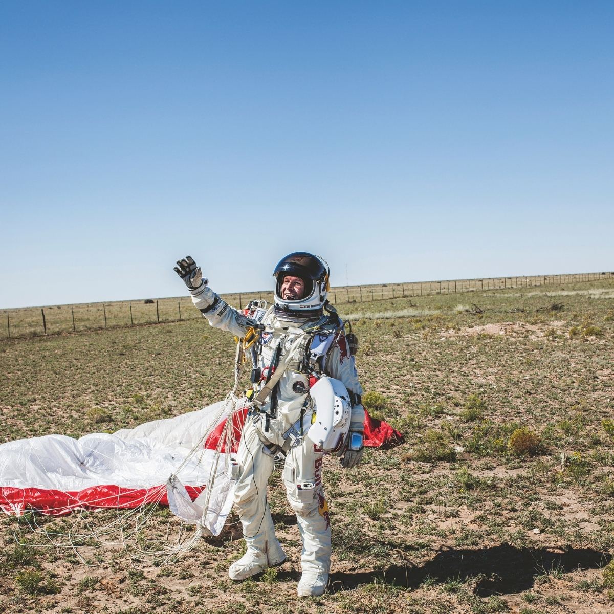 Astronaut in a white space suit stands smiling on a barren field, waving. A parachute lies on the ground, reflecting a triumphant, adventurous mood.