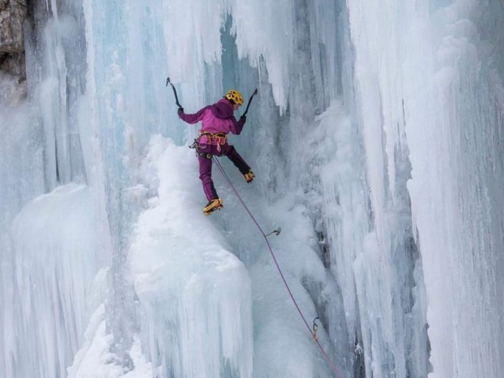 A climber in purple gear, yellow helmet, and ice tools scales a vertical icy wall. The scene conveys bravery and concentration amidst frozen cascades.