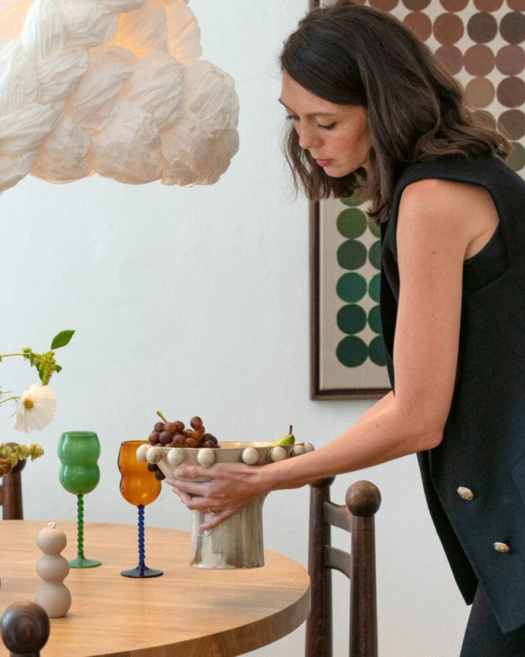 A woman arranges fruit in a decorative bowl on a wooden table. The setting is artistic.