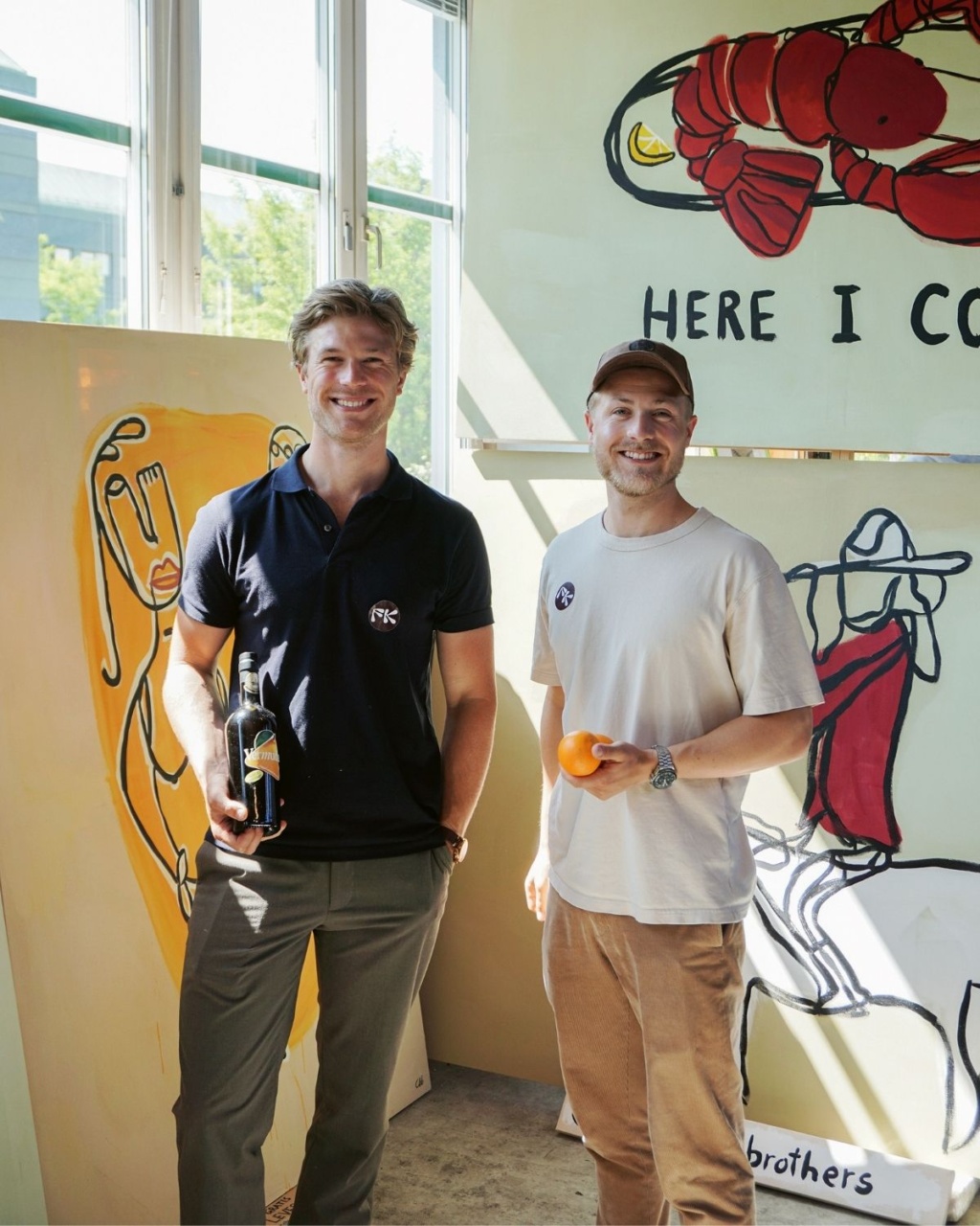 Two men stand smiling in a bright room filled with colorful art. One holds a wine bottle, the other an orange.