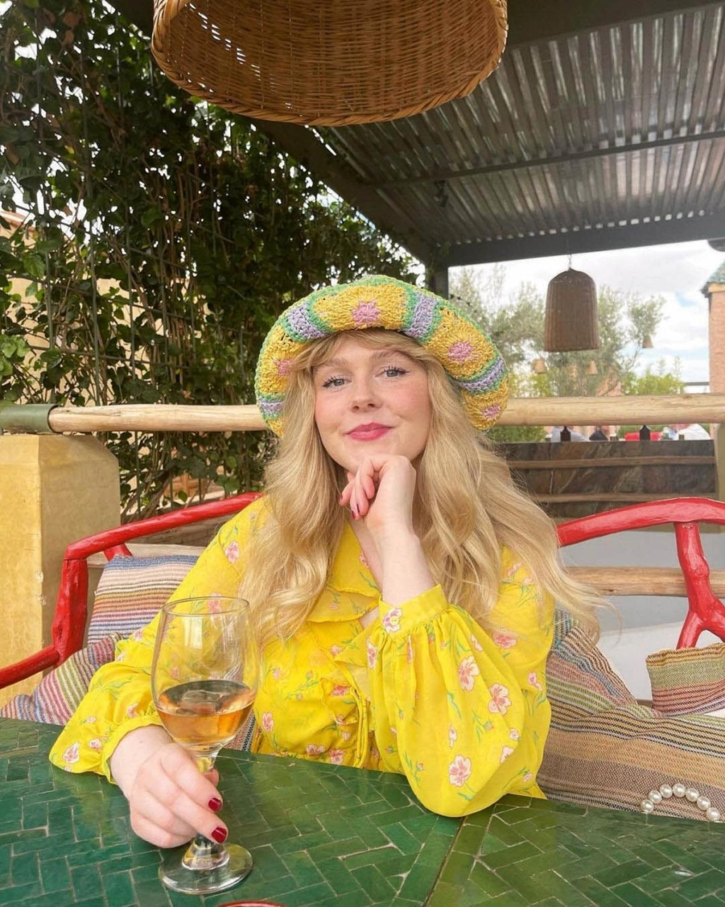 A woman in a yellow floral dress and a colorful crocheted hat sits at a green table outdoors, holding a wine glass and smiling warmly.