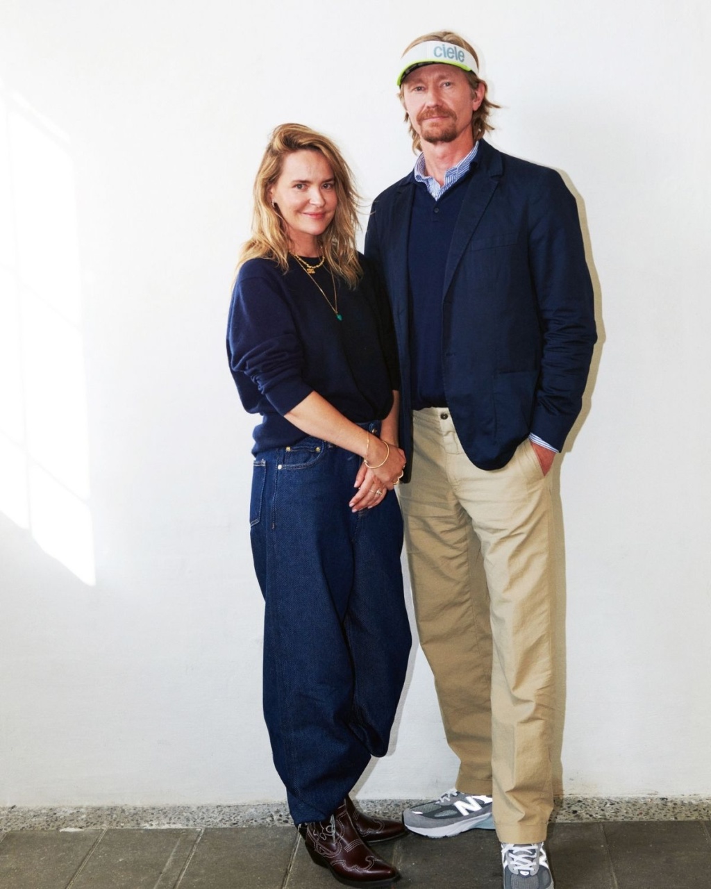 A woman and man stand side by side against a white wall. She's in a navy sweater and jeans; he's in a navy blazer and khakis with a visor.