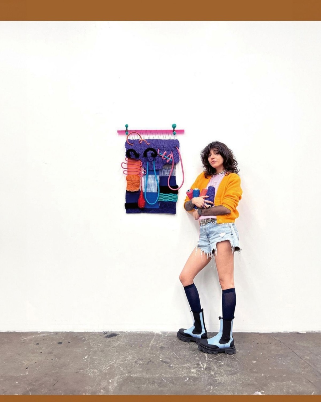 Woman with curly hair stands confidently beside a colorful woven wall hanging. She wears a bright orange sweater, denim shorts, and blue boots.
