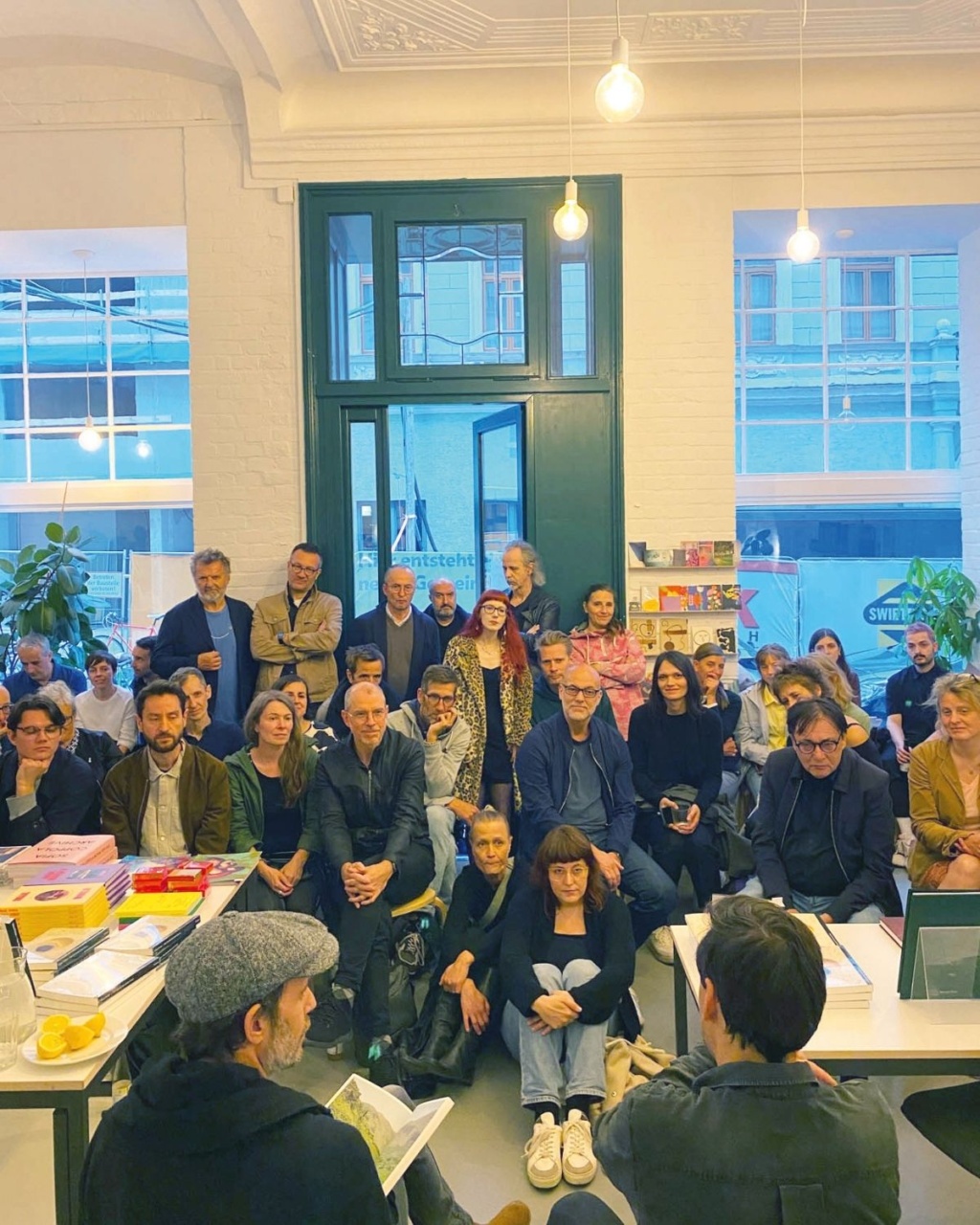 A diverse group of people attentively sits and stands in a well-lit, modern room with large windows.