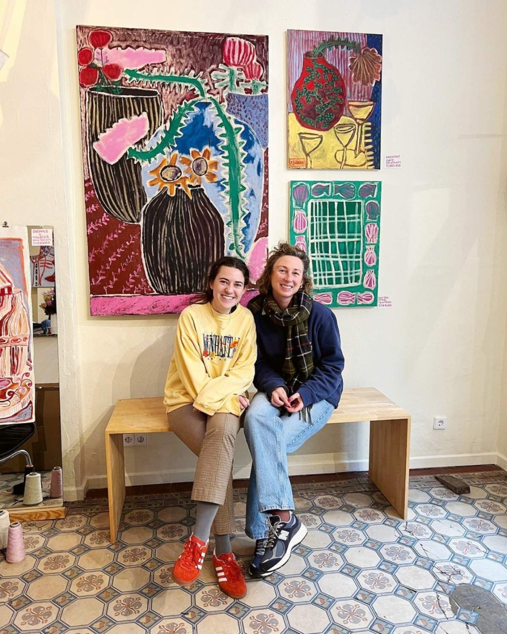 Two people sit on a wooden bench smiling, in front of vibrant abstract paintings. The room has patterned tile flooring.
