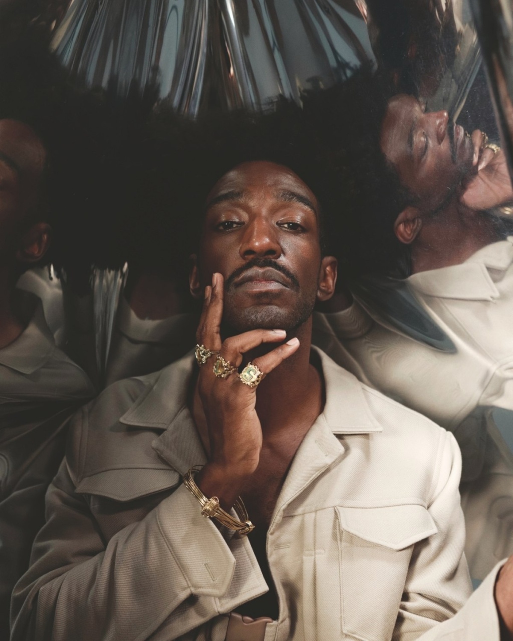 A person poses with their hand to their mouth, showcasing multiple gold rings against a reflective background.