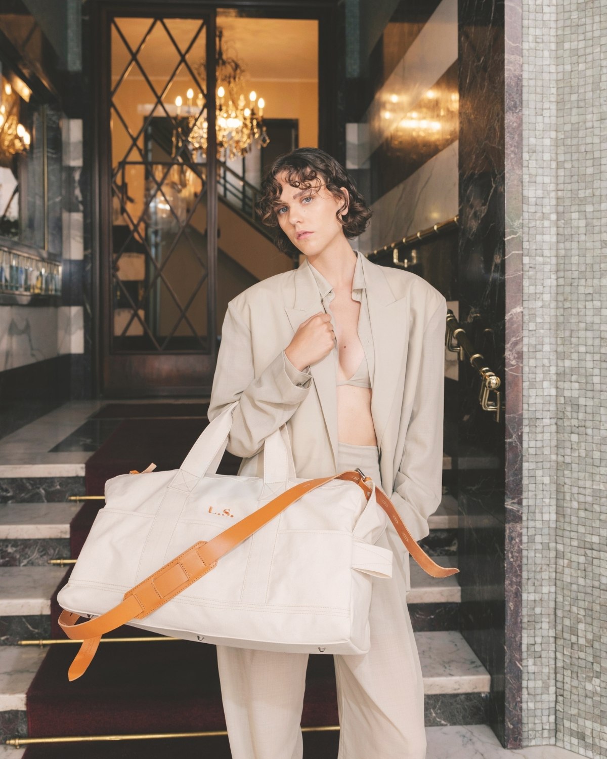 A person in a light beige suit holds a stylish white bag with tan strap in an elegant interior with marble and chandeliers.