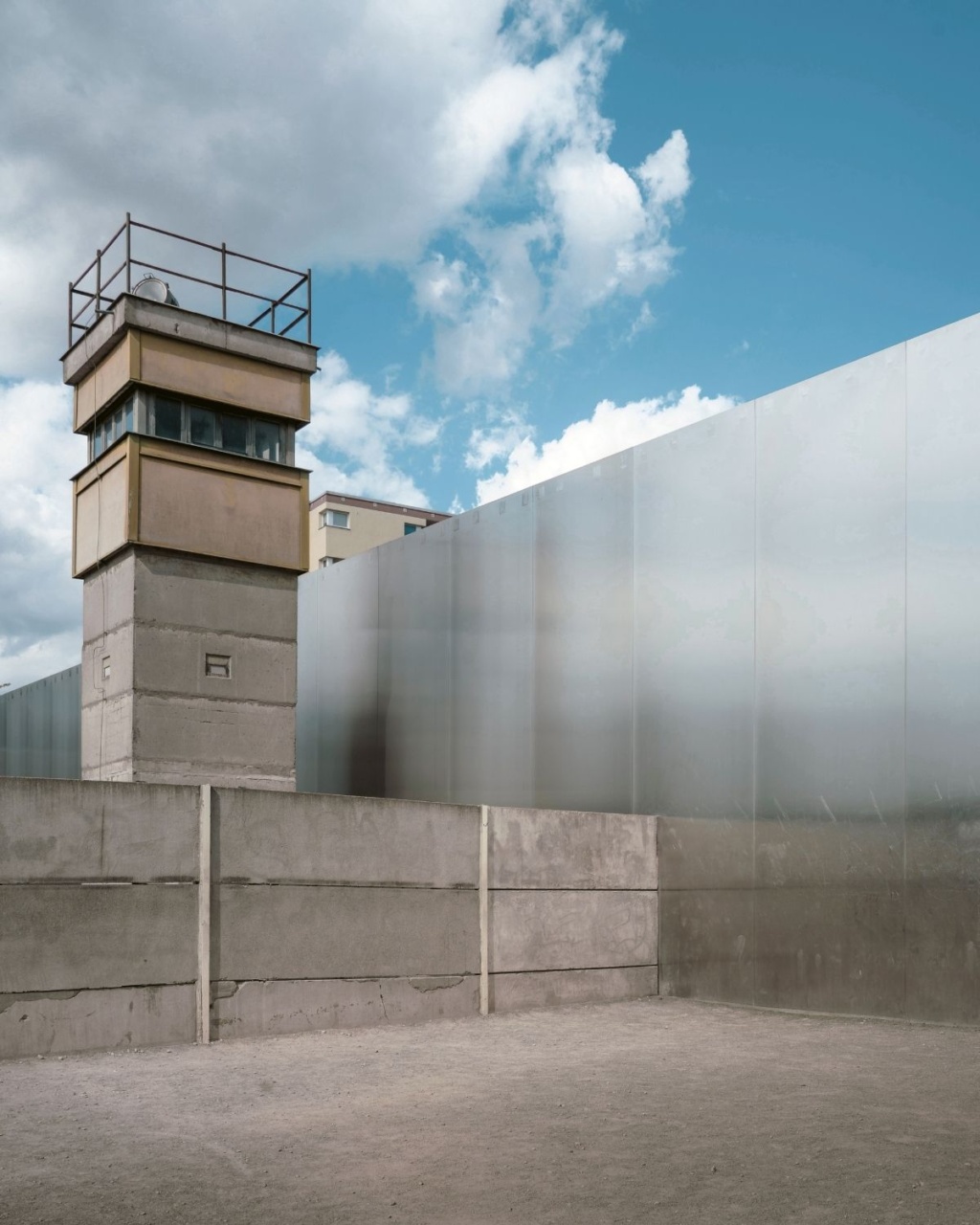 A concrete watchtower stands beside tall metal walls beneath a partly cloudy blue sky, evoking a sense of isolation and surveillance.