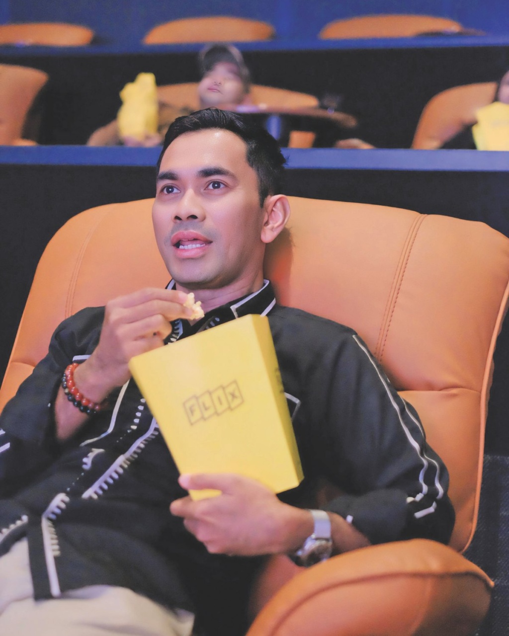 Man in a comfortable theater seat, holding a yellow popcorn box, appears engaged and smiling. Background shows others with similar boxes.