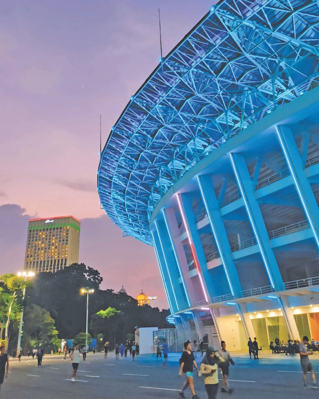 A modern stadium with blue neon lights curves elegantly against a purple sunset sky. People walk below, conveying a lively and vibrant evening scene.
