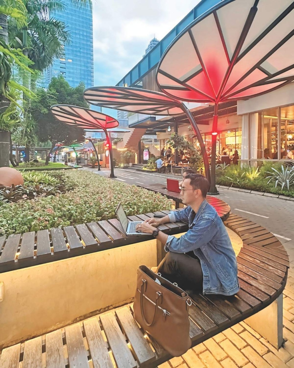 A person in a denim jacket sits on a curved wooden bench using a laptop. Large decorative umbrellas and cityscape are in the background, creating a modern urban atmosphere.