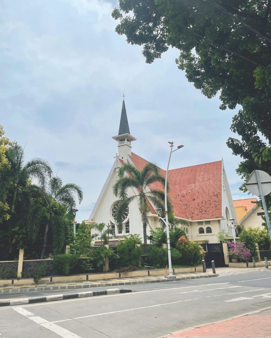A charming church with a tall steeple and red-tiled roof, surrounded by lush palm trees and vibrant greenery, set against a cloudy sky. The street is quiet.