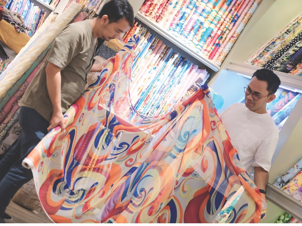 Two men examine a colorful, swirly-patterned fabric in a store. Shelves of vibrant textiles are in the background, creating a lively atmosphere.